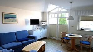 Ein Wohnzimmer mit blauem Sofa, Couchtisch aus Holz, Fernseher, Mikrowelle und einer Essecke mit Stühlen und Tisch.
