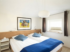 A hotel room with a bed, a bedside table, a framed picture on the wall, a window with curtains, and a hanging light fixture.