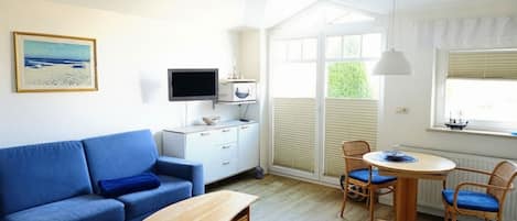 A modern living room with a blue sofa, a wooden coffee table, a TV, a dining table with chairs, and a window with blinds.