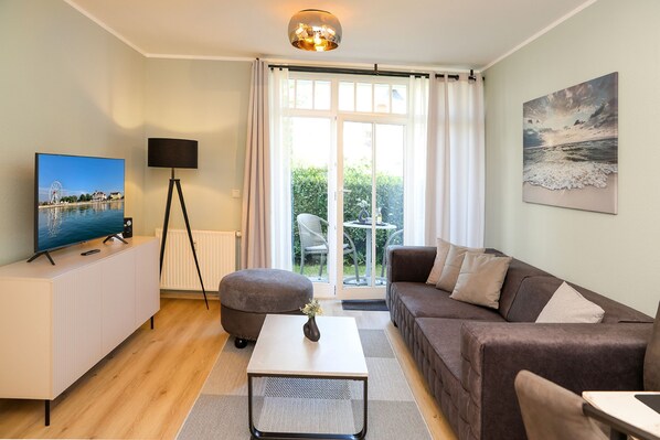 A modern living room with a television, a floor lamp, a sofa, a coffee table, and a view of a garden through a glass door.