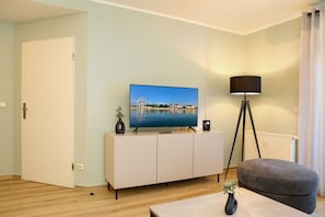 A living room with a television on a cabinet, a floor lamp, a sofa, and a small table with a vase. - Rosenhof 17 Apartment - Unit 17 (Kühlungsborn)
