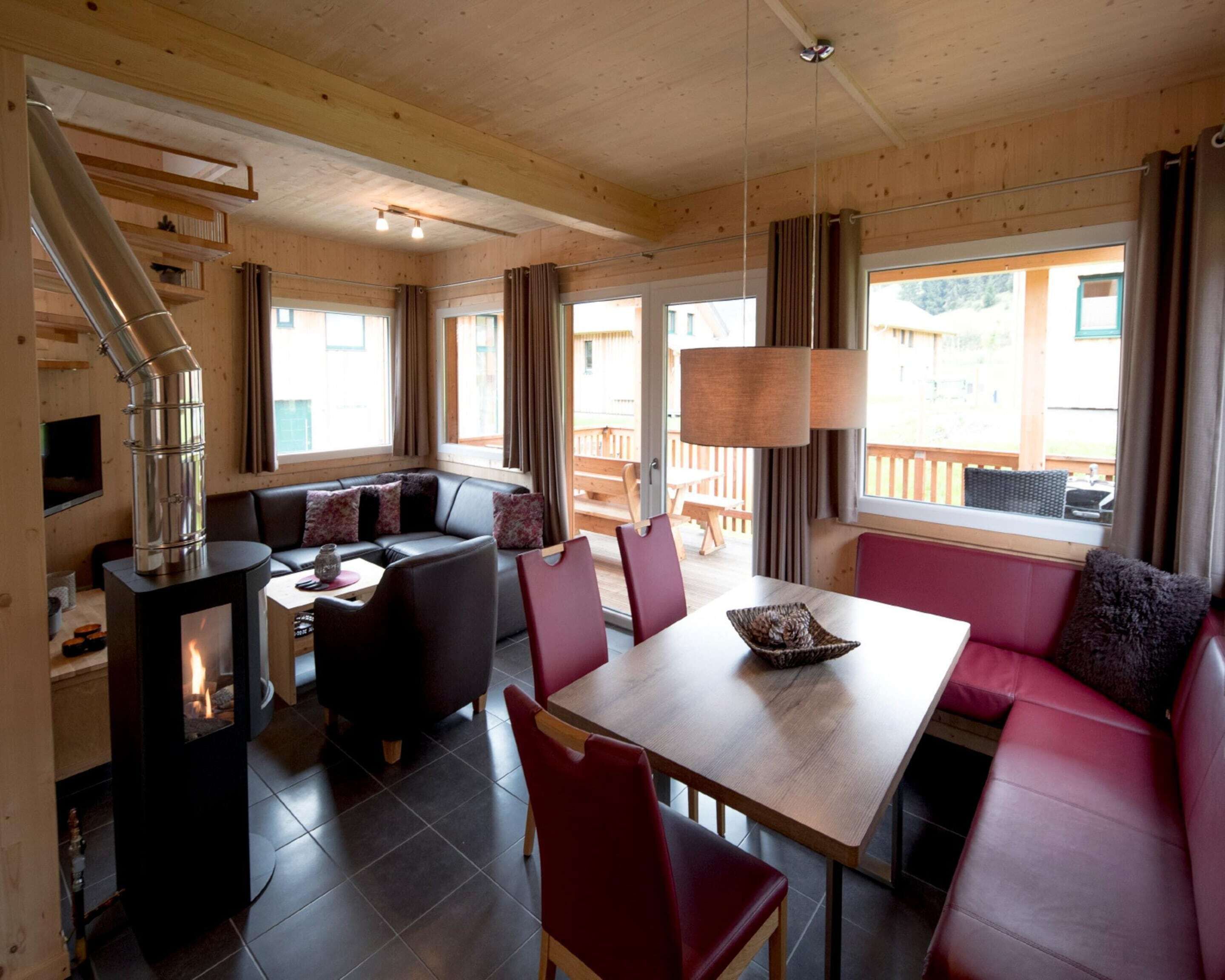 Interior acogedor de una cabaña con chimenea, paredes de madera, zona de comedor y área de descanso con vistas al exterior.