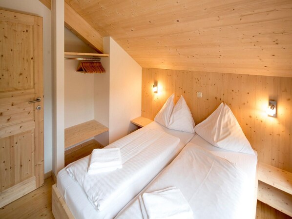 A wooden-paneled room with two beds, a wooden wardrobe, and a shelf with folded clothes.