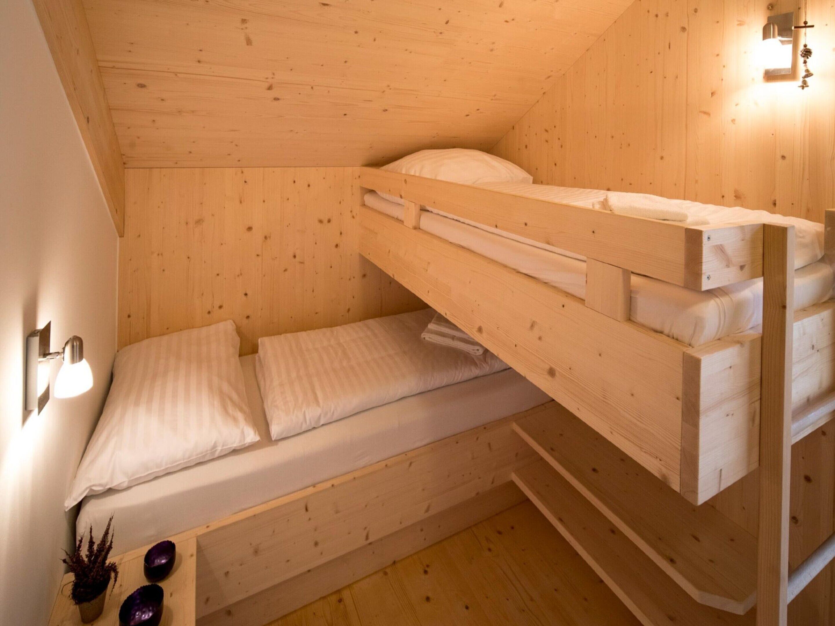 Une chambre avec un lit superposé en bois, un plafond en pente, une lampe fixée au mur et une petite étagère ornée d’objets décoratifs.