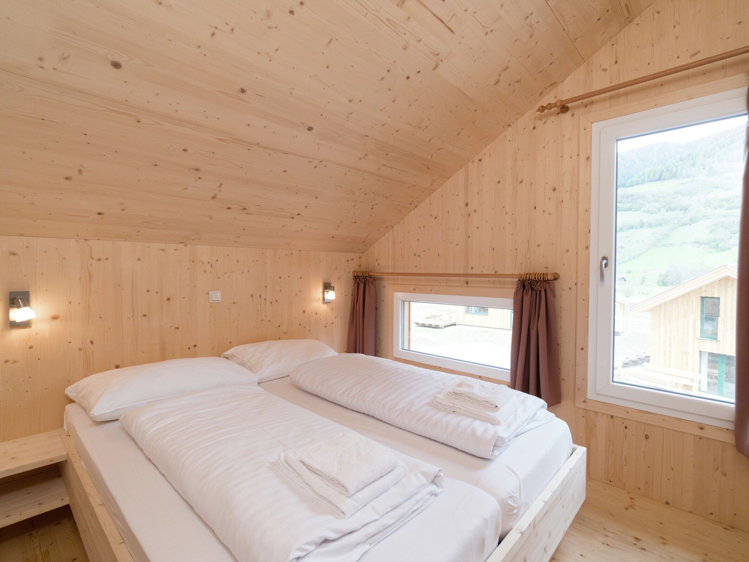 A wooden cabin room with two beds, a window with curtains, and a small wall-mounted light.