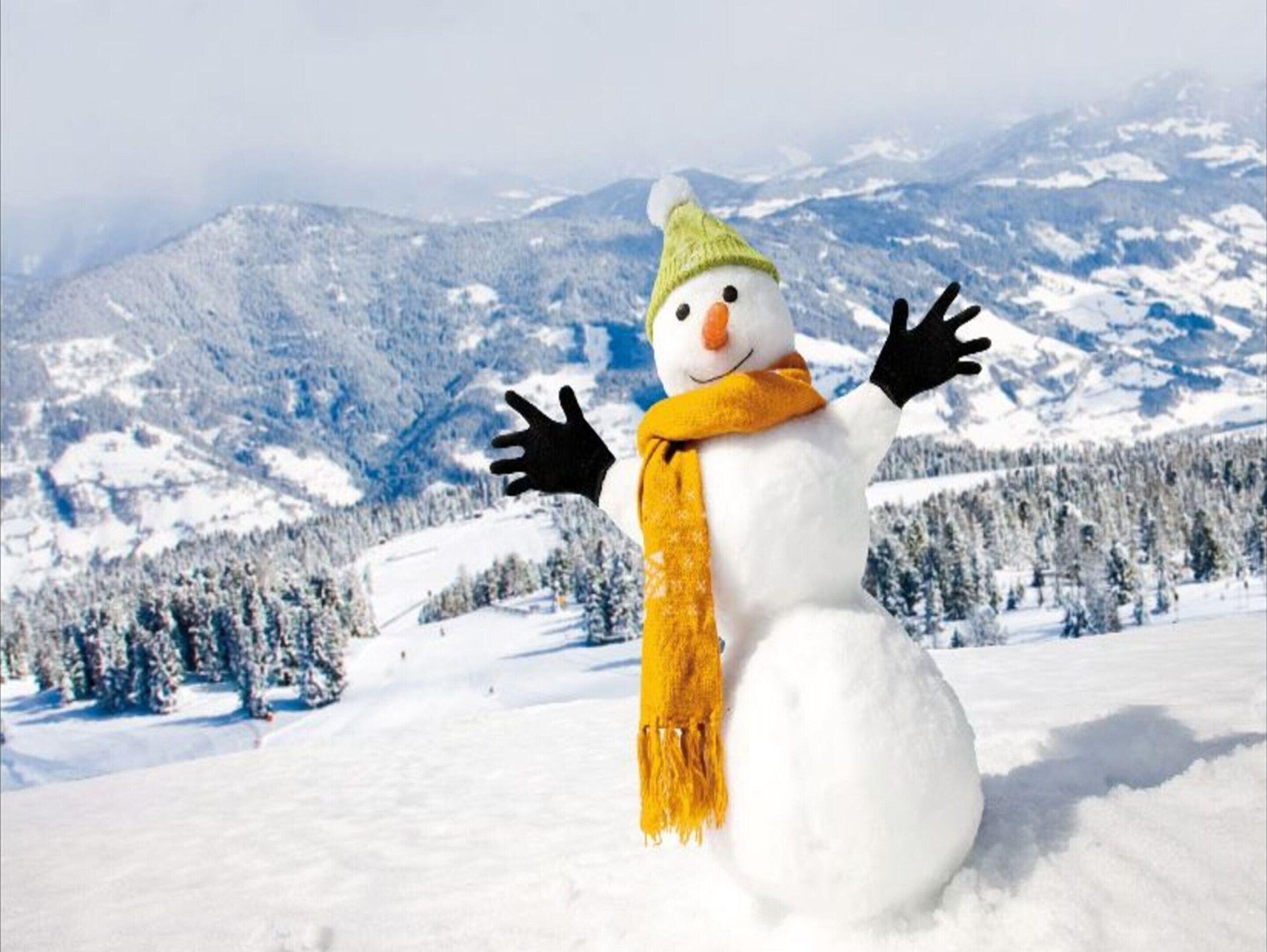 A snowman with a green hat and yellow scarf in a snowy landscape with mountains in the background.