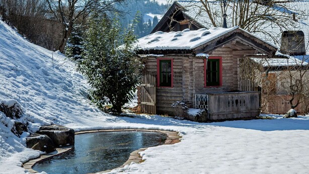 A wooden cabin with a snow-covered roof, a small pond, and a mountainous backdrop. - Huge Holiday Home in Hopfgarten im Brixental near Ski Lift (Hopfgarten im Brixental)
