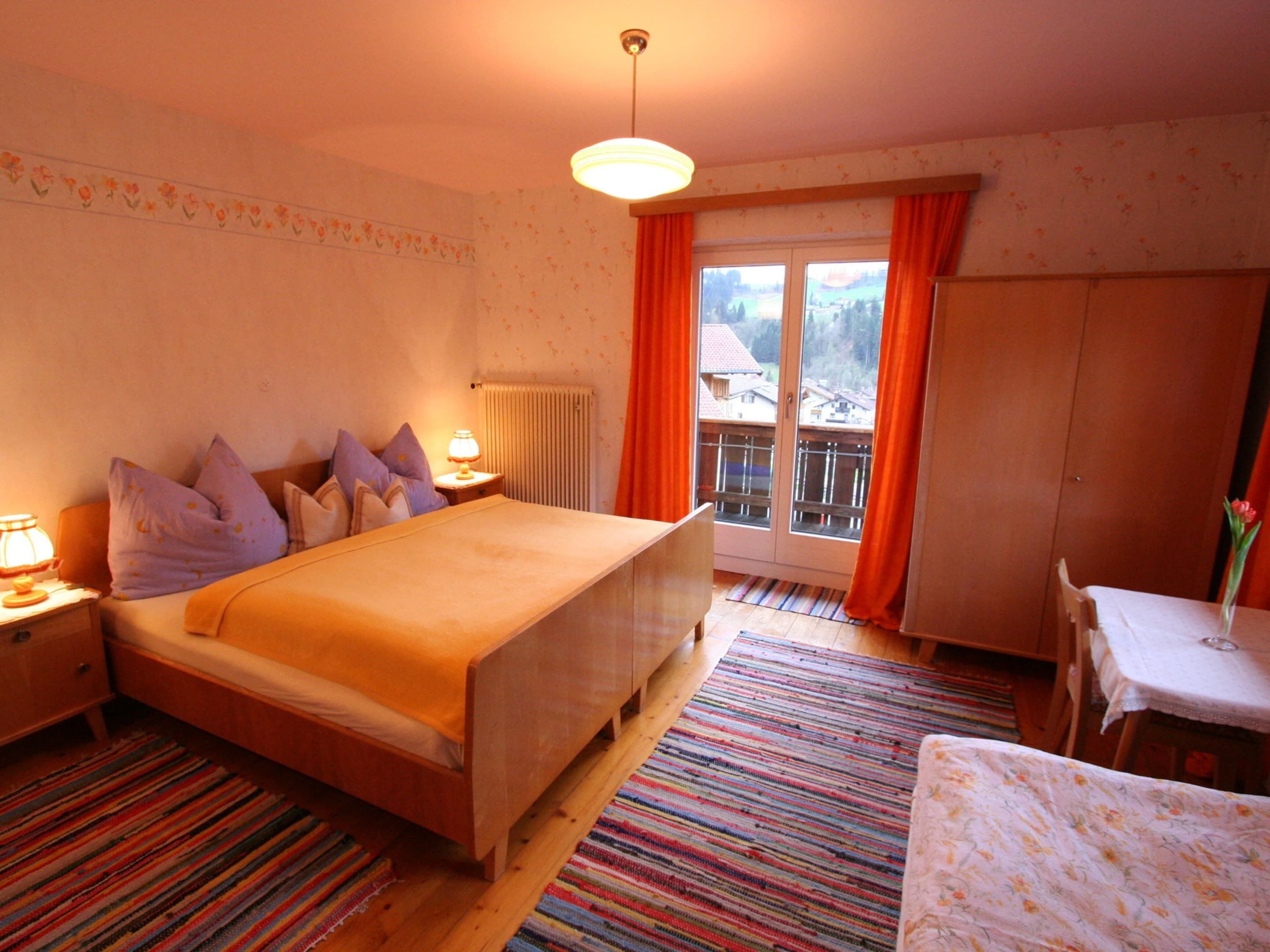 A bedroom with a bed, a nightstand, a lamp, a window with curtains, a radiator, a wooden floor, and a small table with a chair.