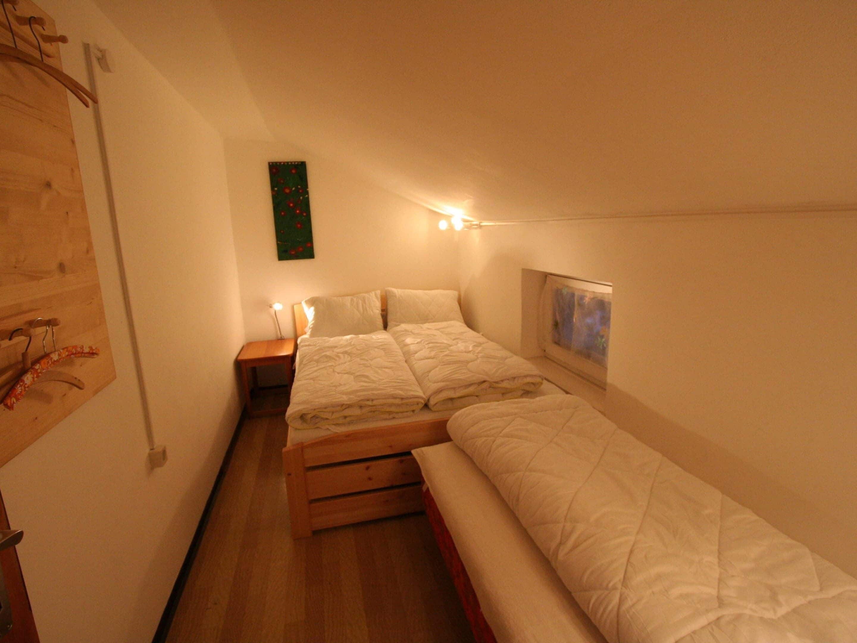 A small, cozy bedroom with a single bed, a nightstand, a wall-mounted shelf, and a small window.
