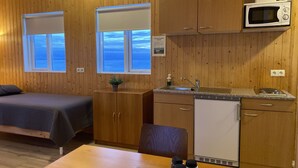 A compact kitchen with wooden cabinets, a microwave, a sink, and a small refrigerator. There are two windows providing views of the outside. - Beindalsholt (Rangárþing ytra)