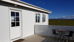 A white building with a wooden deck, a door, and windows. There is a view of mountains in the background. - Beindalsholt (Rangárþing ytra)