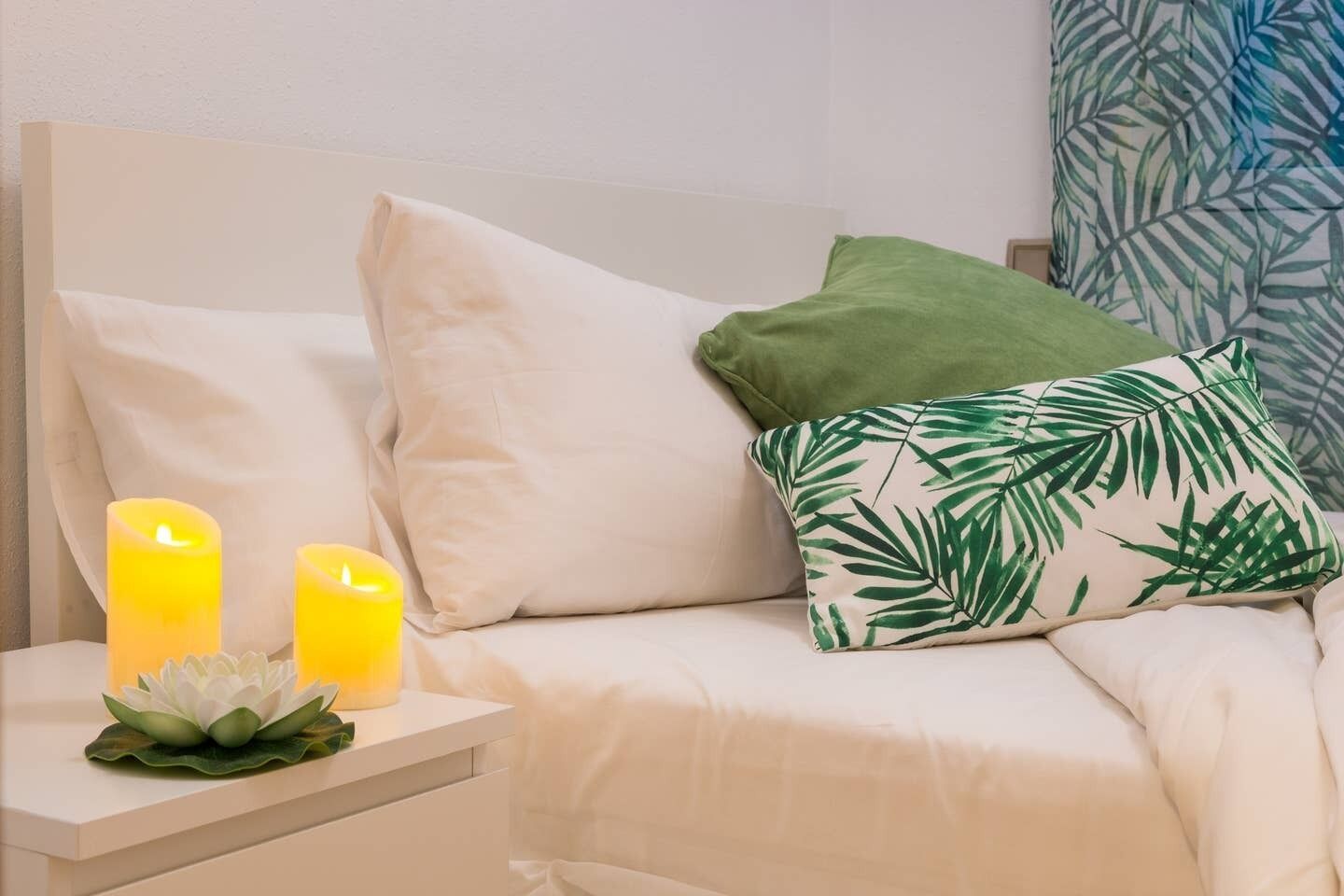A bed with white bedding, two lit candles, and a decorative plant on a bedside table.