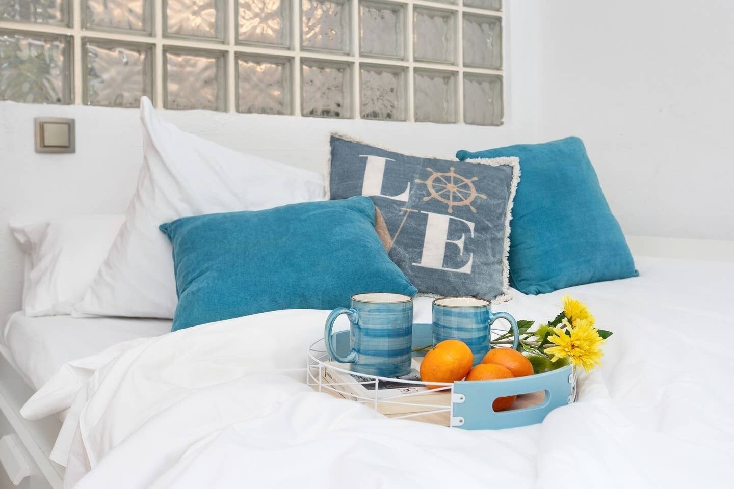 Ein Bett mit weißer Bettwäsche, blauen Kissen und einem Tablett mit zwei Tassen und Orangen.