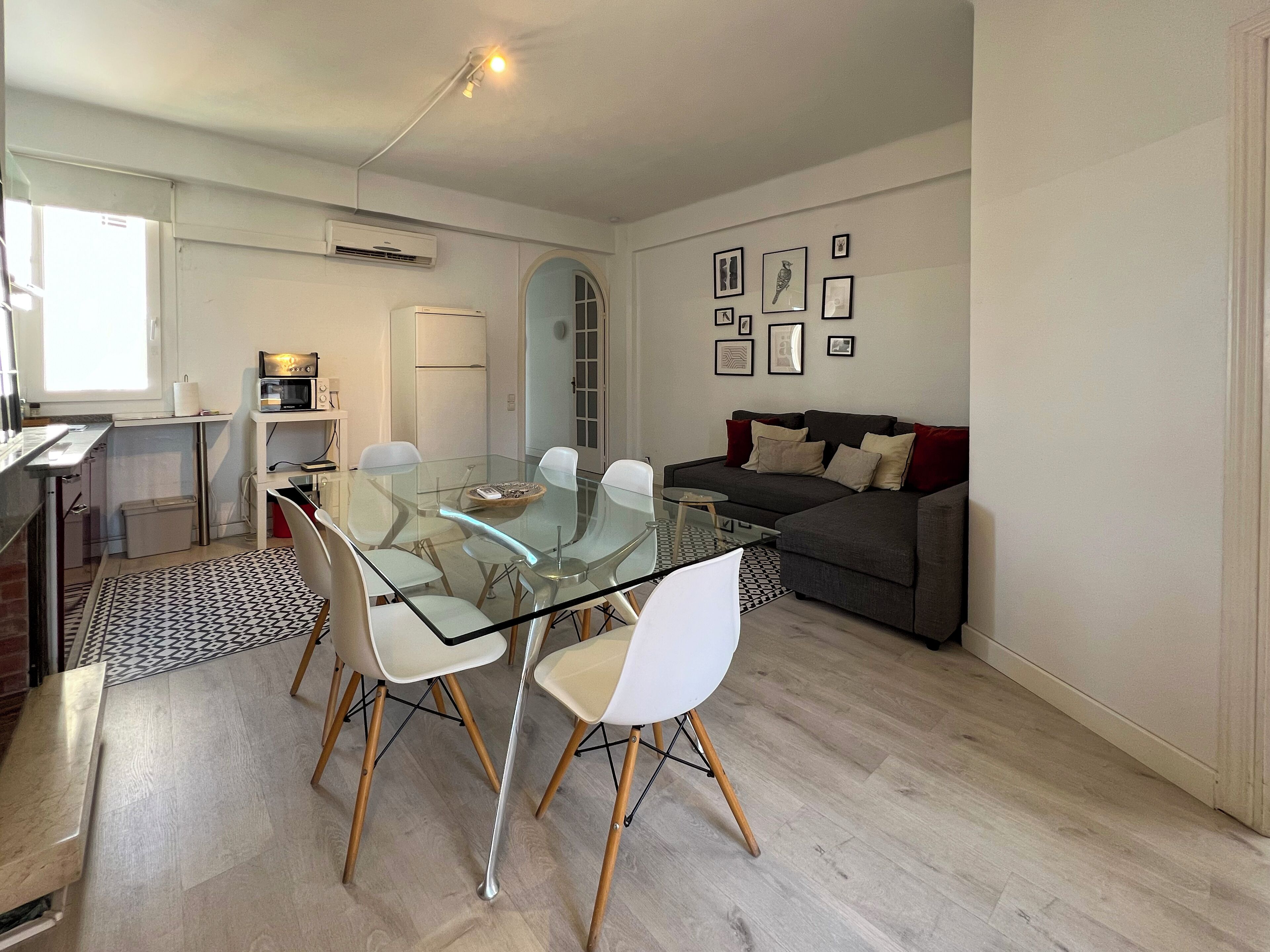 Un salon moderne avec une table à manger en verre entourée de chaises, un canapé gris avec des coussins et un coin cuisine doté de meubles blancs et d’un climatiseur.