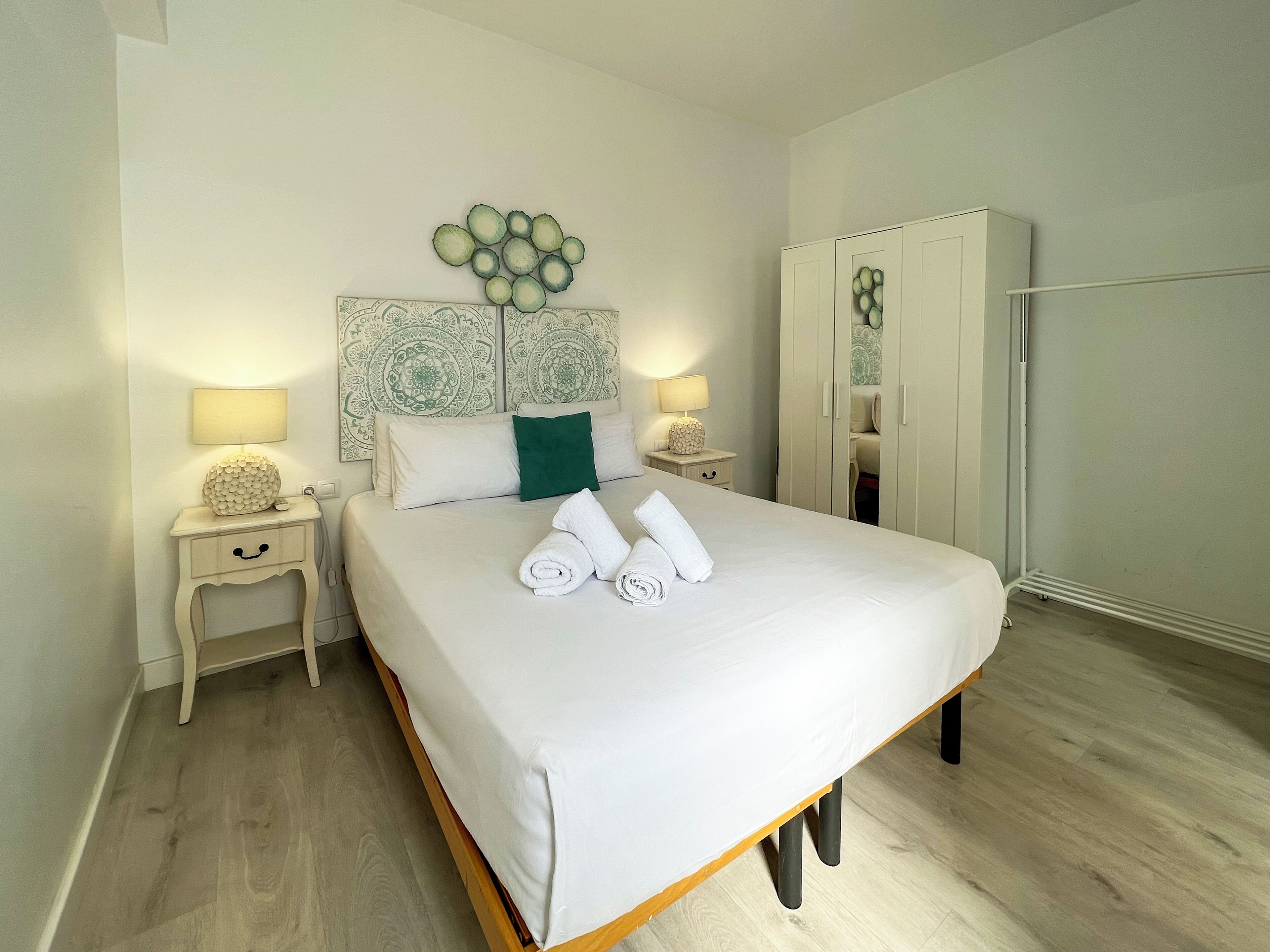 A neatly made bed with white linens and rolled towels, a bedside table with a lamp, and a wardrobe with a mirror.