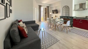 A modern living room with a grey sofa, a dining table with chairs, a kitchen area with red cabinets, and a TV mounted on the wall. - Stay U-nique Apartment Sèneca (Barcelona)