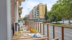 A balcony with a wooden table, a bottle of wine, two wine glasses, a bowl of fruit, and a magazine. - Stay U-nique Apartment Sicília (Barcelona)