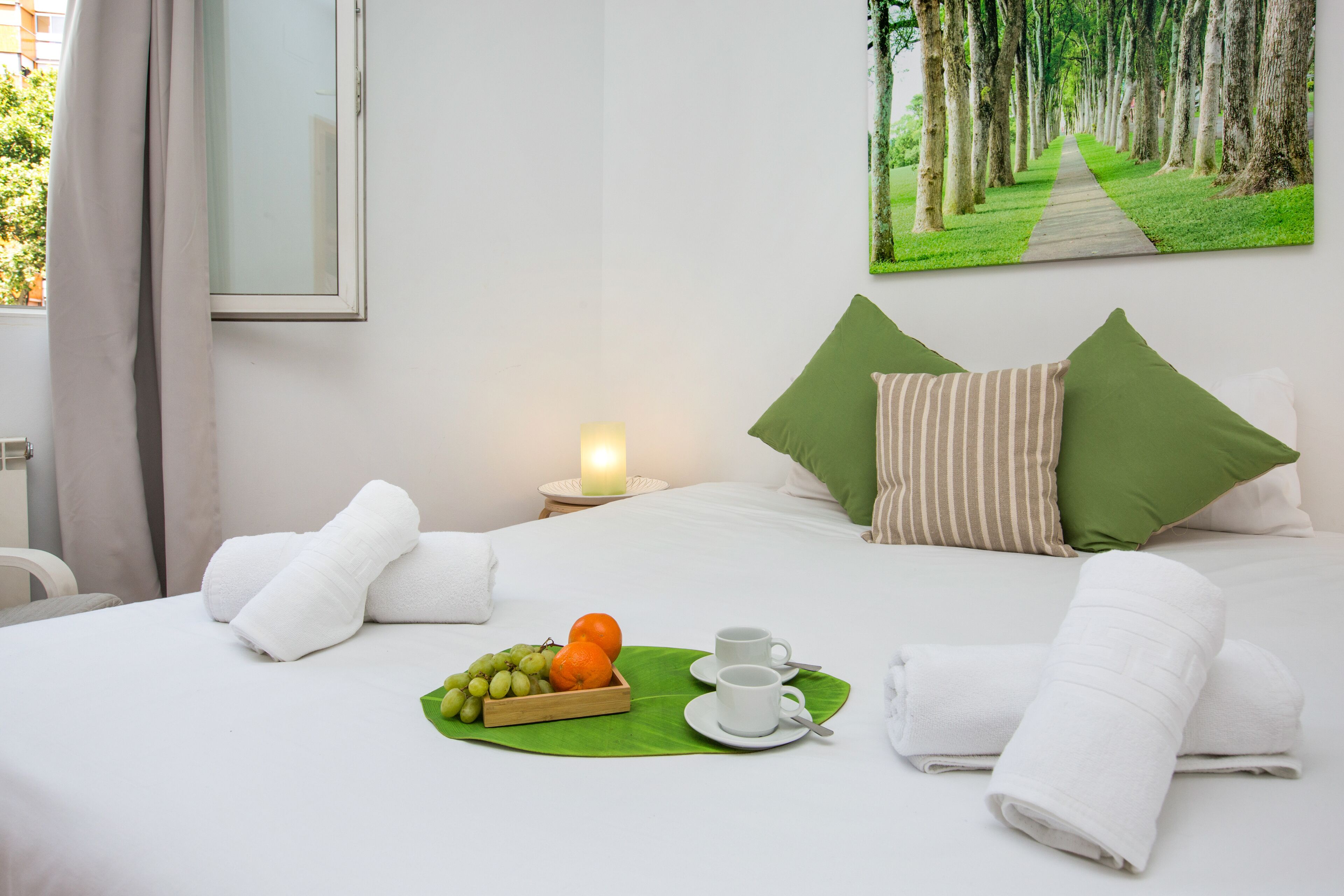 A neatly made bed with white linens, green pillows, and a fruit basket with grapes and oranges.