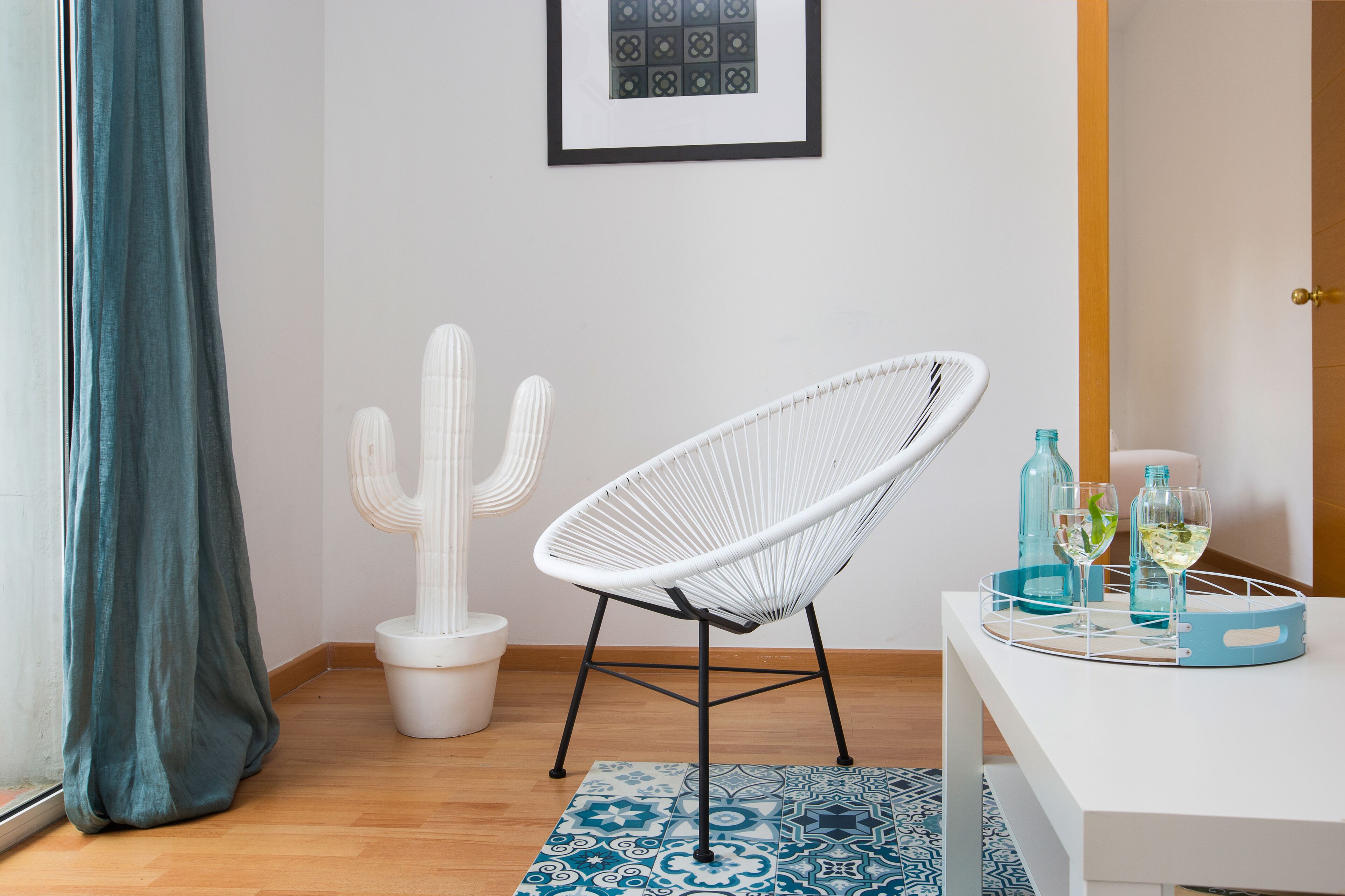 Una habitación con una silla blanca, una escultura de cactus, un cuadro en la pared y una mesa con dos vasos y botellas.