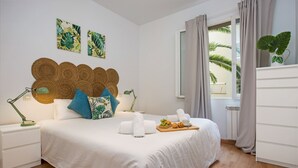 A neatly made bed with white linens, a tray of fruits and a cup, a nightstand with a lamp, and a view of palm trees through the window. - Stay U-nique Apartment Sicília (Barcelona)