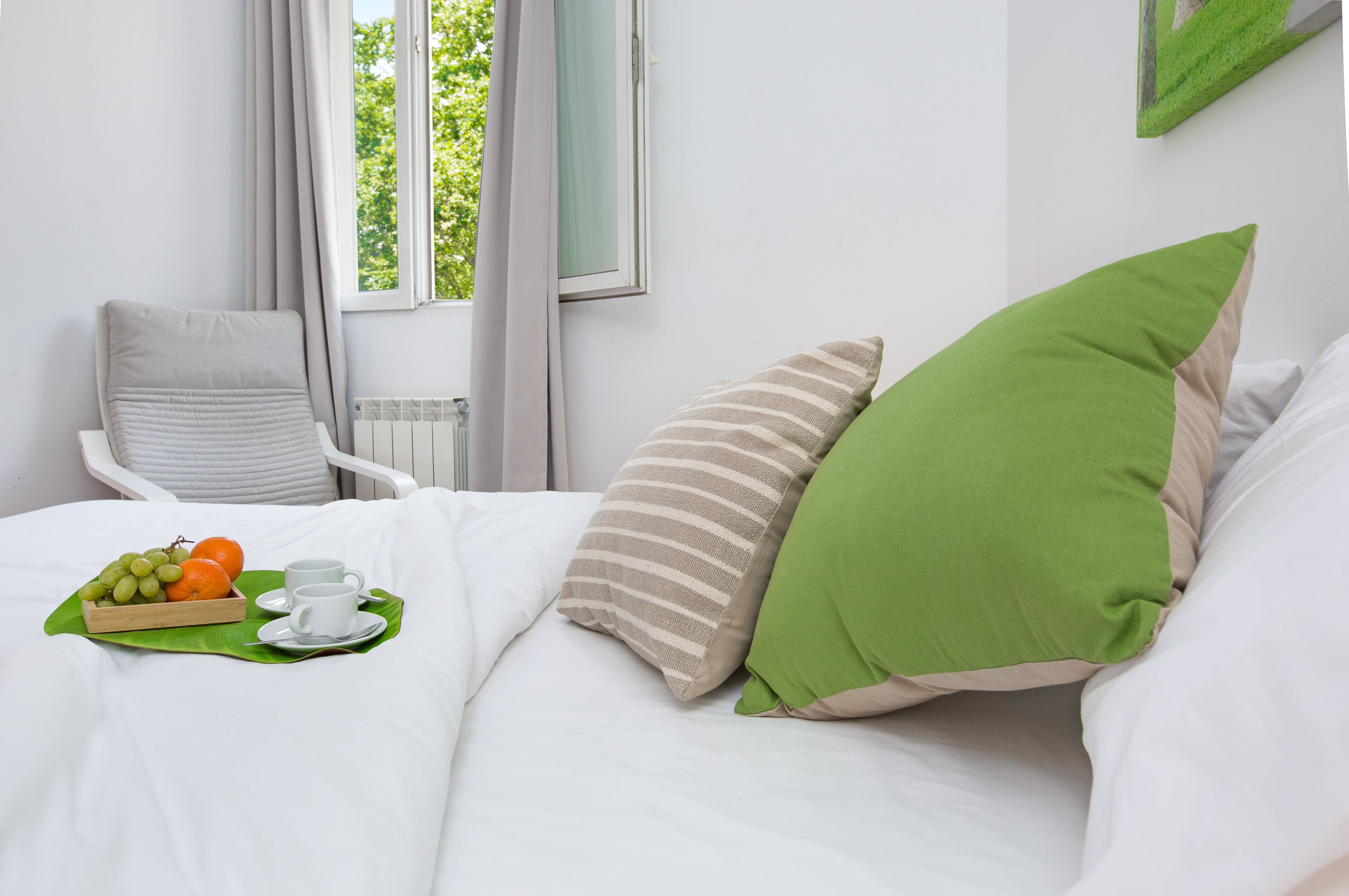 Een net opgemaakt bed met witte lakens, een groen kussen en een gestreept kussen. Er staat een dienblad met fruit en een kopje op het bed.