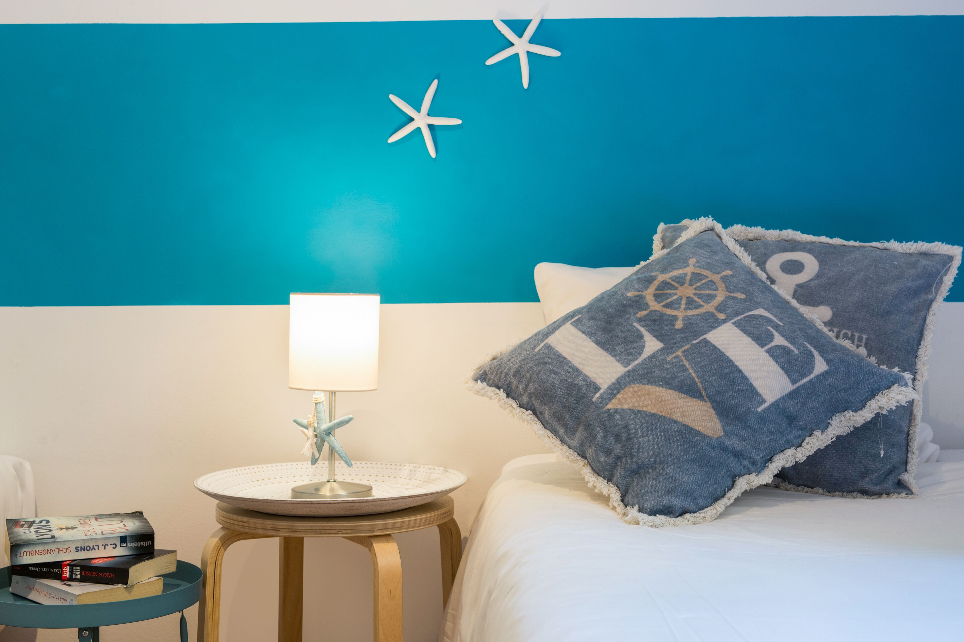En pænt redt seng med en hvid dyne og to puder, hvoraf den ene har et maritimt design. Et lille rundt bord med en lampe og en stjernefiskdekoration står ved siden af sengen. Der er bøger stablet på bordet.