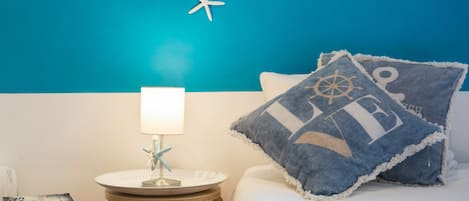 En pænt redt seng med en hvid dyne og to puder, hvoraf den ene har et maritimt design. Et lille rundt bord med en lampe og en stjernefiskdekoration står ved siden af sengen. Der er bøger stablet på bordet.