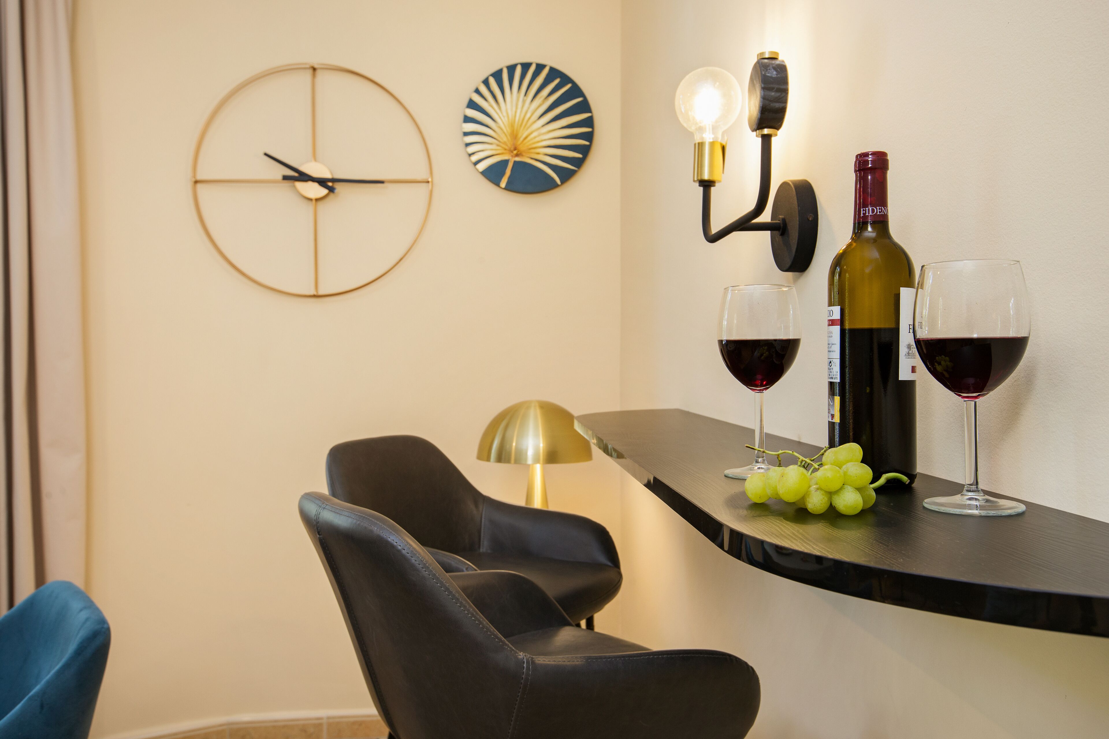 Un comedor moderno con un reloj redondo en la pared, una lámpara fijada a la pared, una botella de vino y dos copas de vino tinto sobre una encimera.