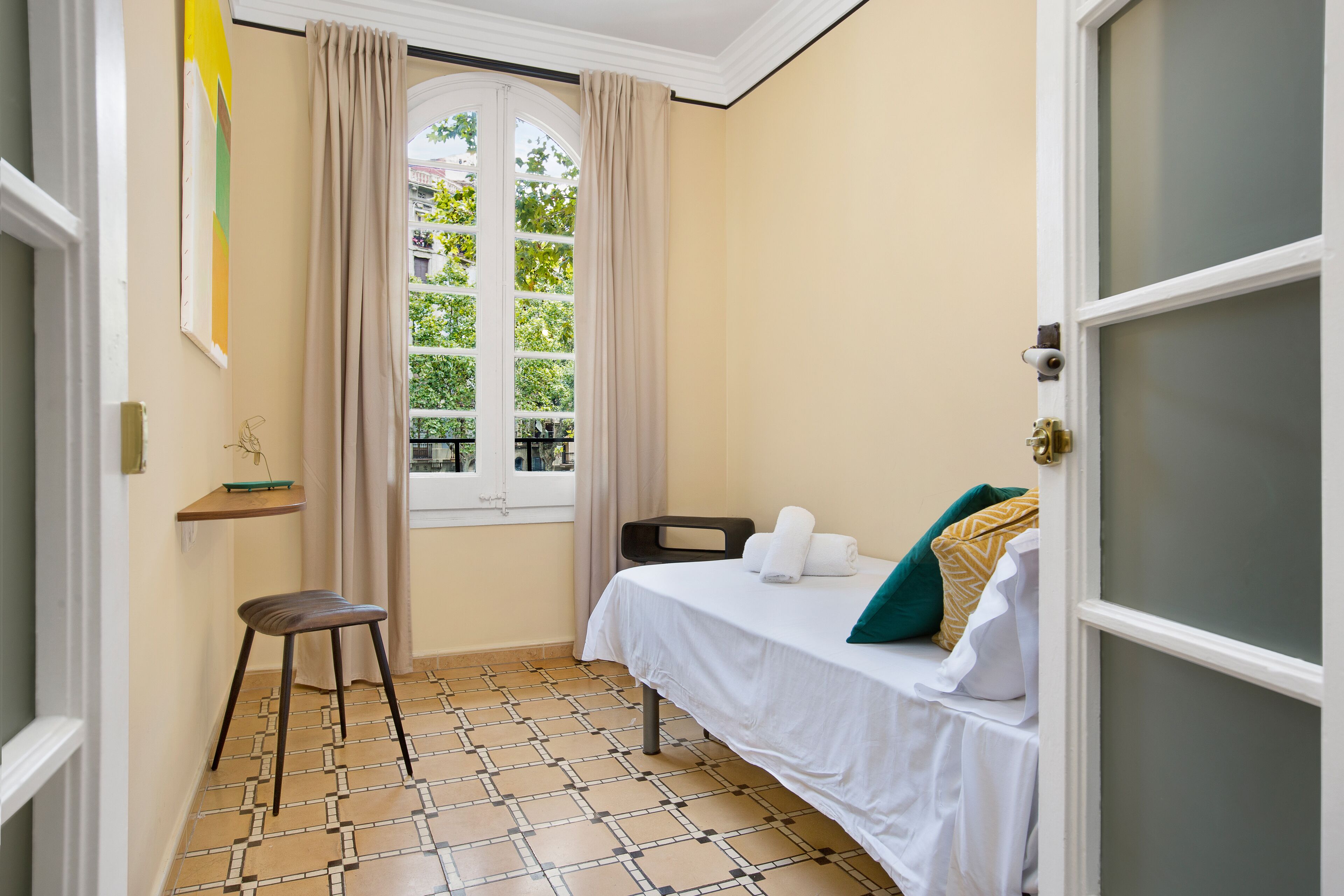 A room with a bed, a chair, a window with curtains, a shelf with a book, and a tiled floor.