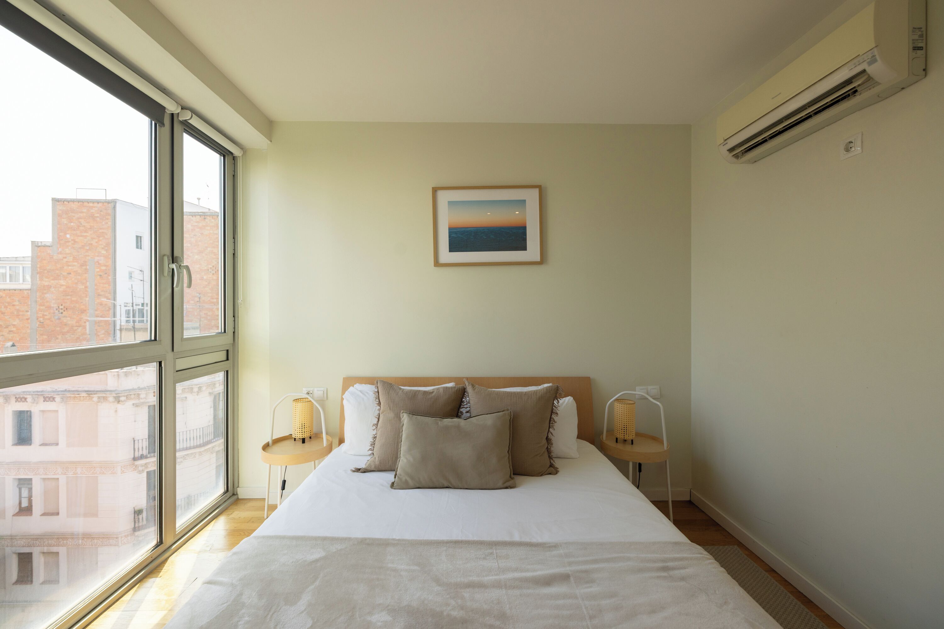A bedroom with a large window, a bed with pillows, a framed picture on the wall, and an air conditioning unit.
