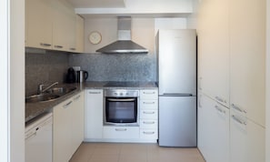 A compact kitchen with a built-in oven, a stainless steel range hood, a refrigerator, and a clock on the wall.