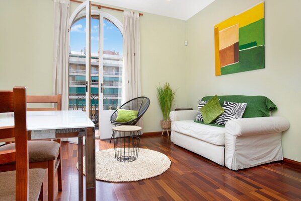 A living room with a dining table, a white sofa, a wicker chair, and a view of a building through a glass door.
