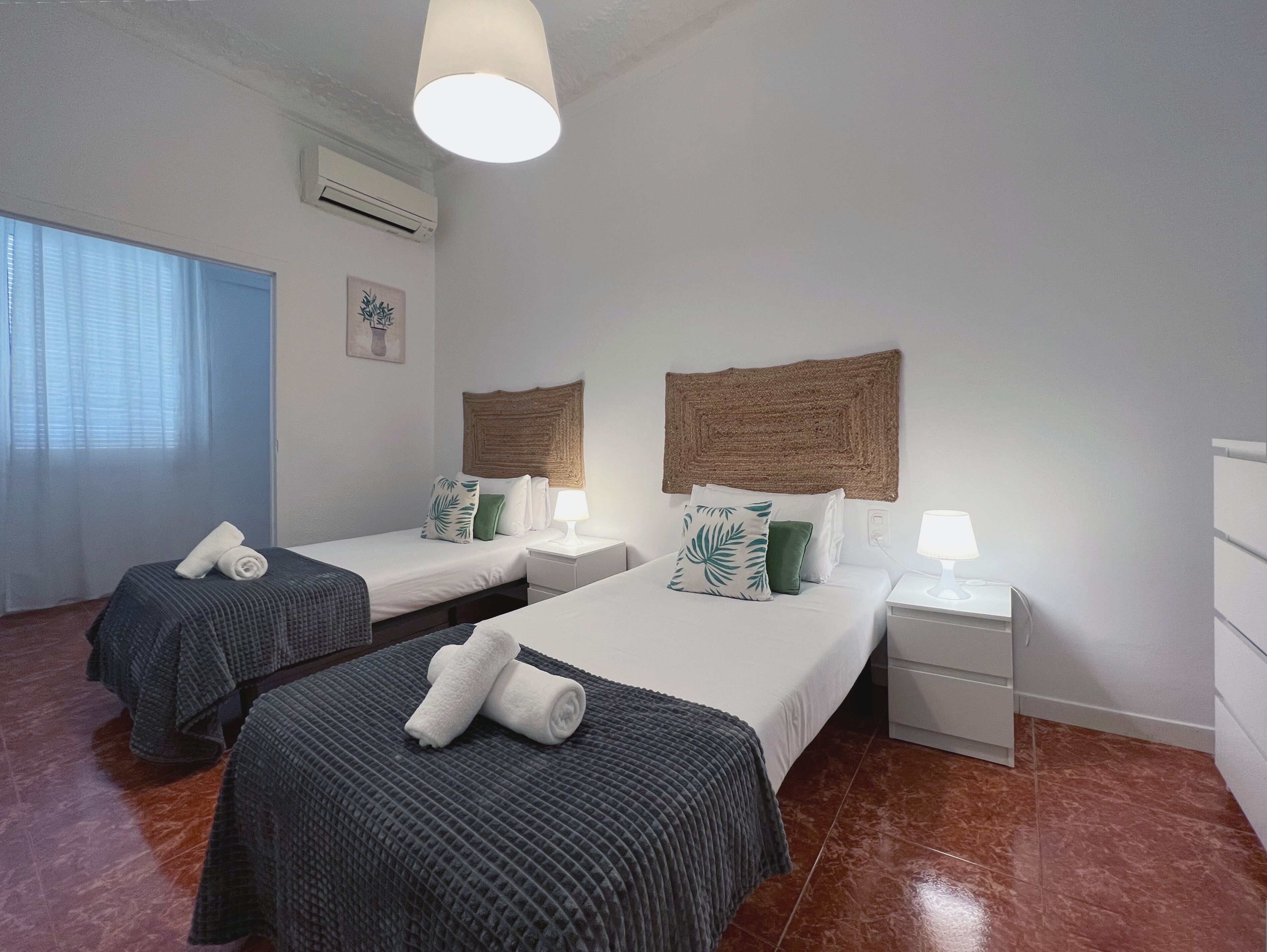 Habitación de hotel con dos camas, cada una con cabecera, mesitas de noche con lámparas, una ventana con cortinas y un equipo de aire acondicionado en la pared.