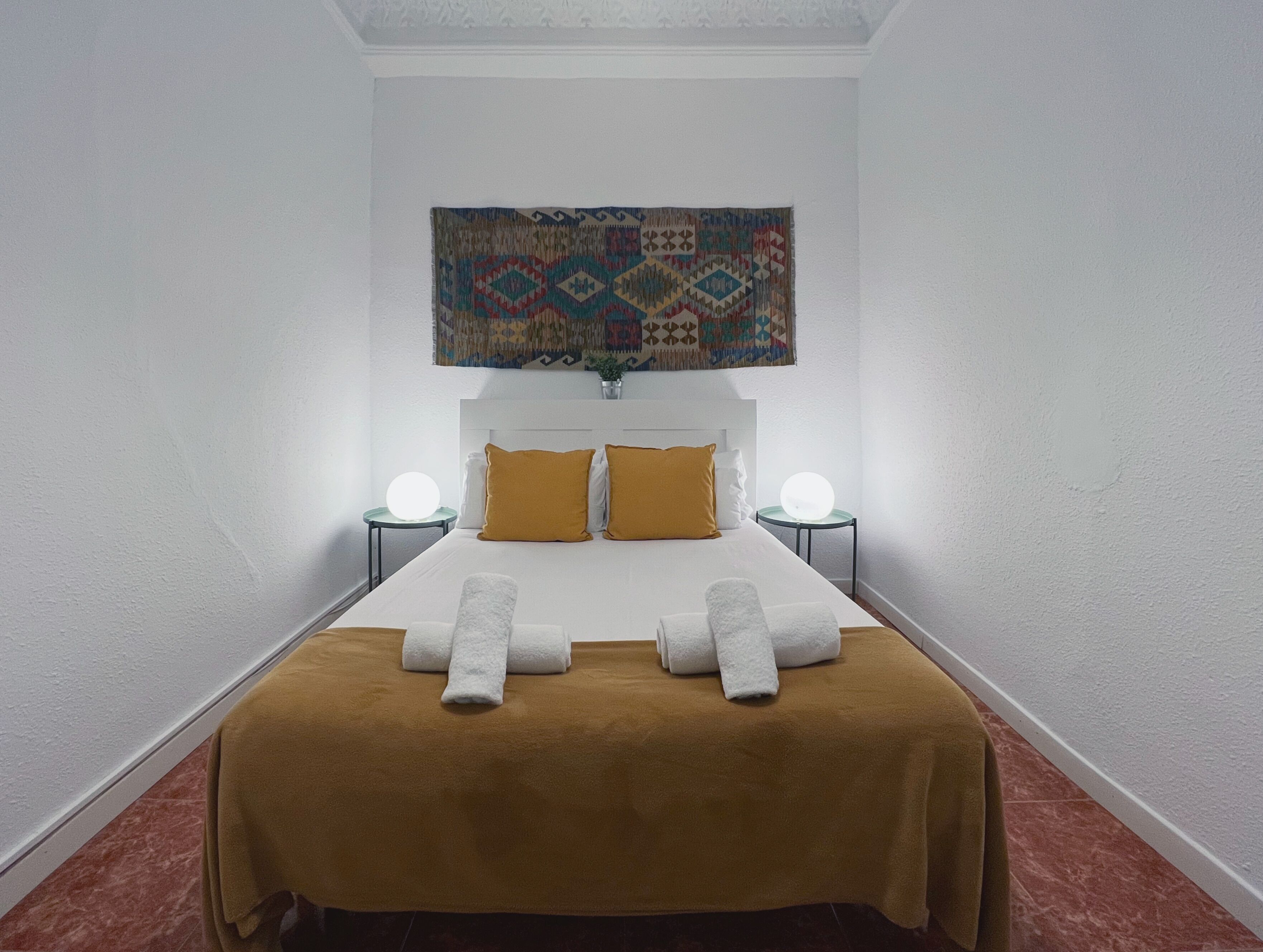 Una habitación de hotel con una cama, dos mesitas de noche, un tapiz en la pared y un suelo de baldosas rojas.