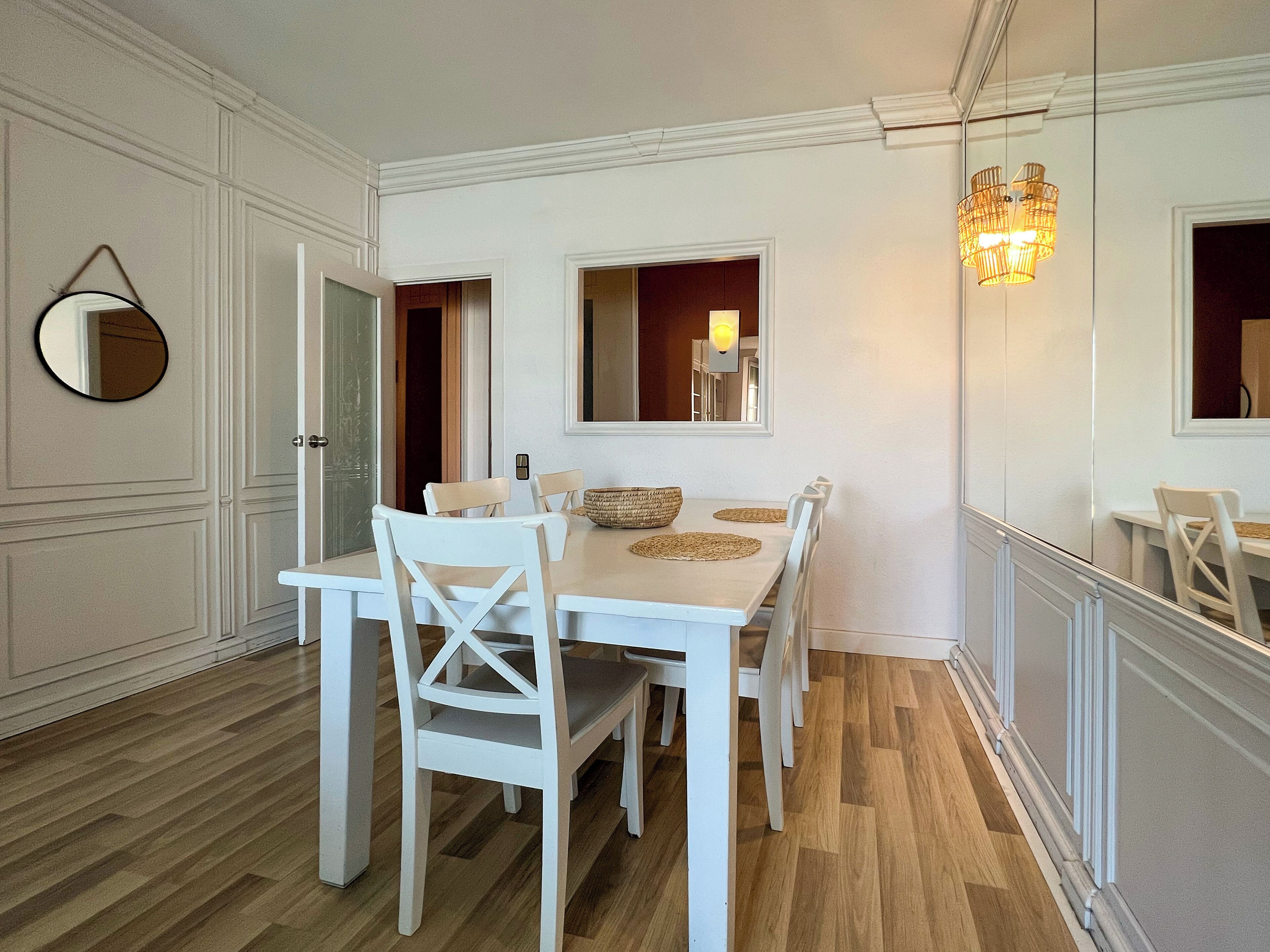 Un coin repas avec une table et des chaises blanches, un miroir et un luminaire fixé au mur.