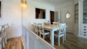 Een eetgedeelte met een witte tafel en stoelen, een kroonluchter en uitzicht op een keuken met witte kasten.