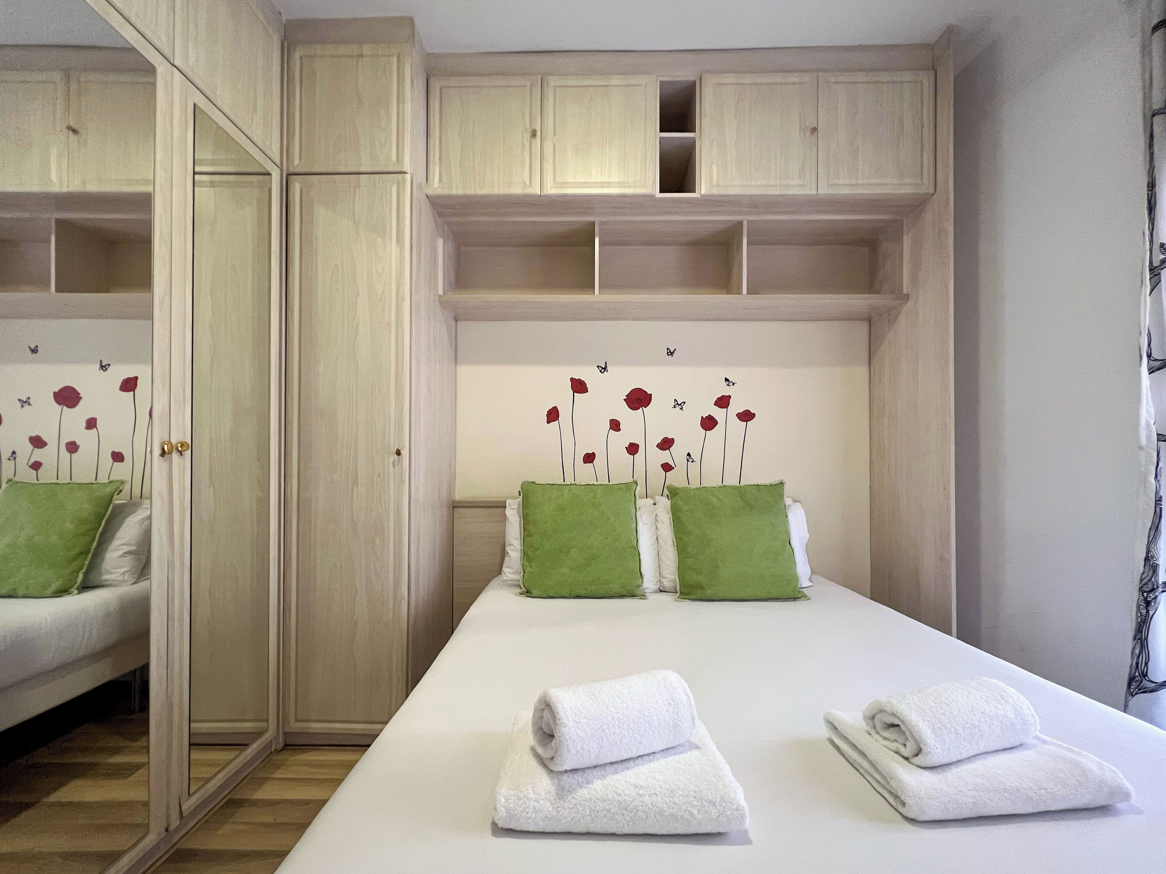 Une chambre d’hôtel avec un lit, deux oreillers verts, deux serviettes blanches, une armoire en bois et un mur orné d’un motif floral.