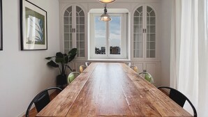 A long wooden dining table with chairs, a hanging light fixture, and large arched windows with white frames.