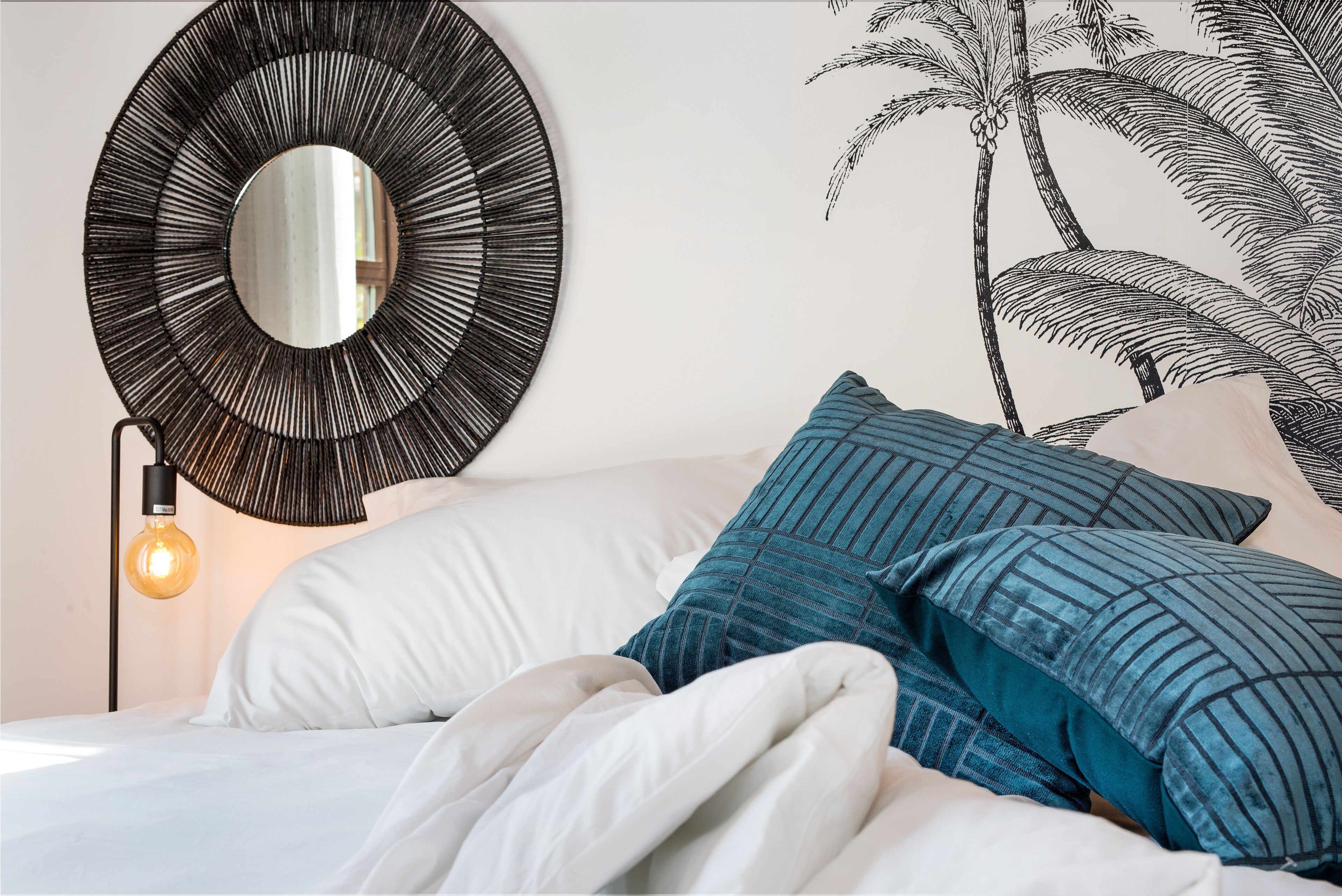 A bed with white bedding and blue striped pillows, a round mirror on the wall, and a wall-mounted light fixture.