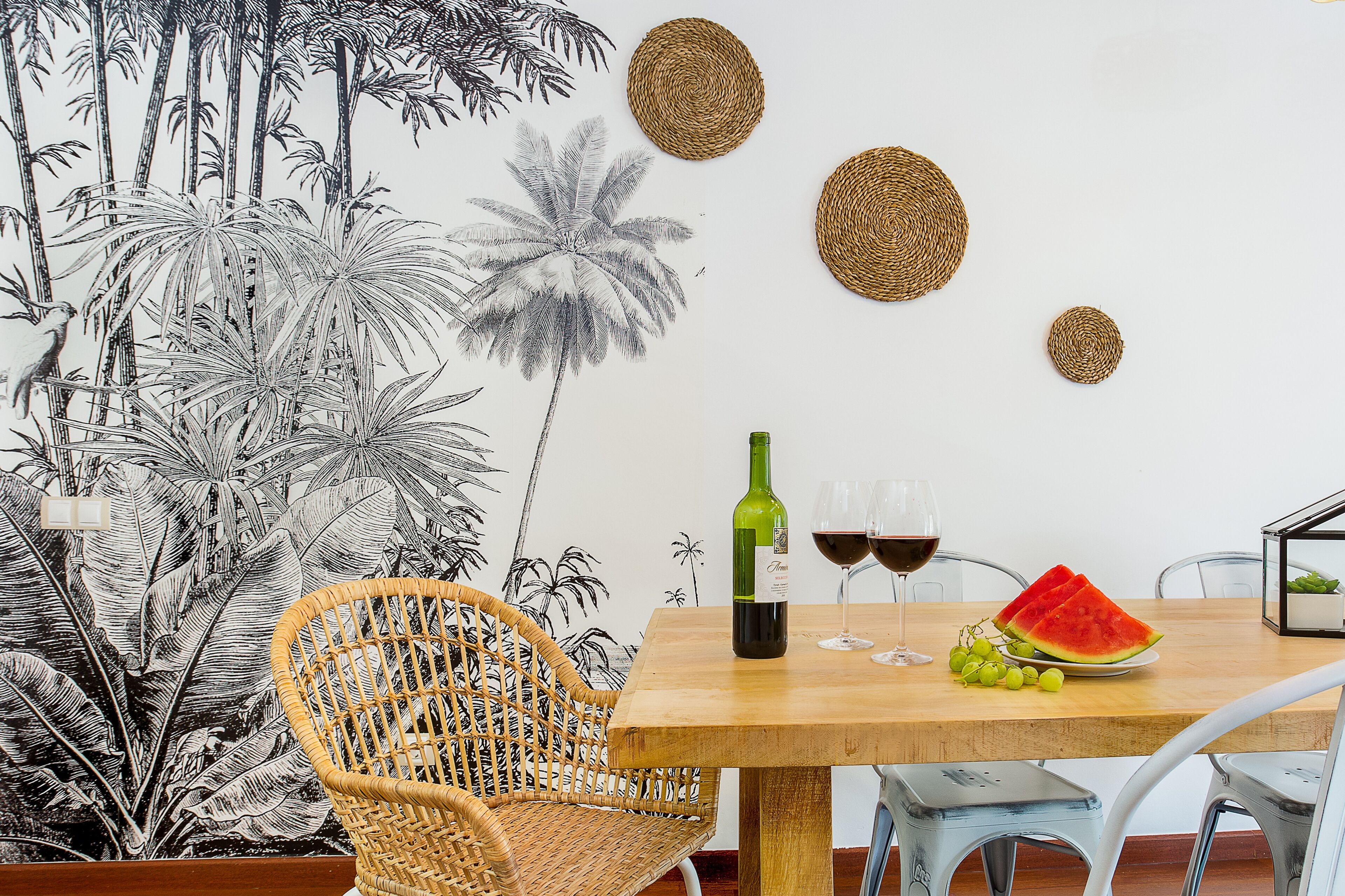 Un coin repas avec une table en bois, des chaises en osier, une bouteille de vin, des verres et une assiette de pastèque et de raisins.