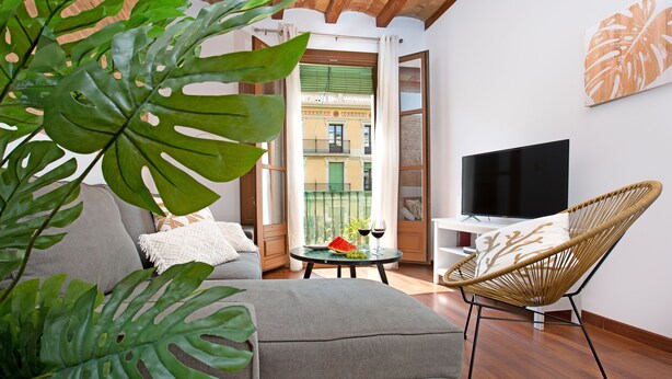 A modern living room with a sofa, a wicker chair, a TV, and a balcony with a view of buildings. - Stay U-nique Apartments Girona (Barcelona)