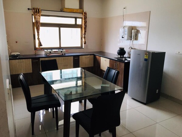 Une cuisine compacte avec une table à manger en verre, des chaises noires, un réfrigérateur et une fenêtre avec des rideaux.