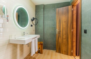 Un baño moderno con un lavamanos blanco, un espejo redondo, una puerta de madera y una zona de ducha.
