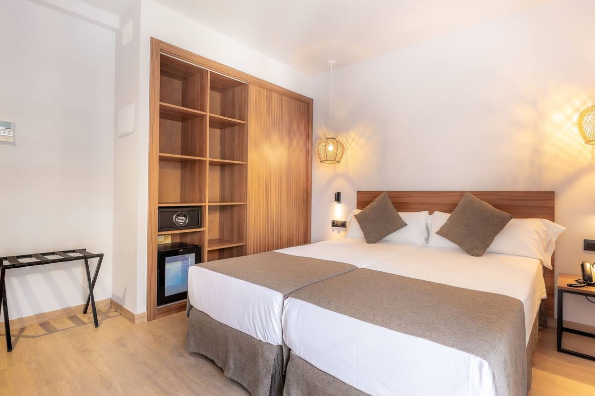 A hotel room with a large bed, bedside lamps, a wardrobe, a small table, and a minibar.