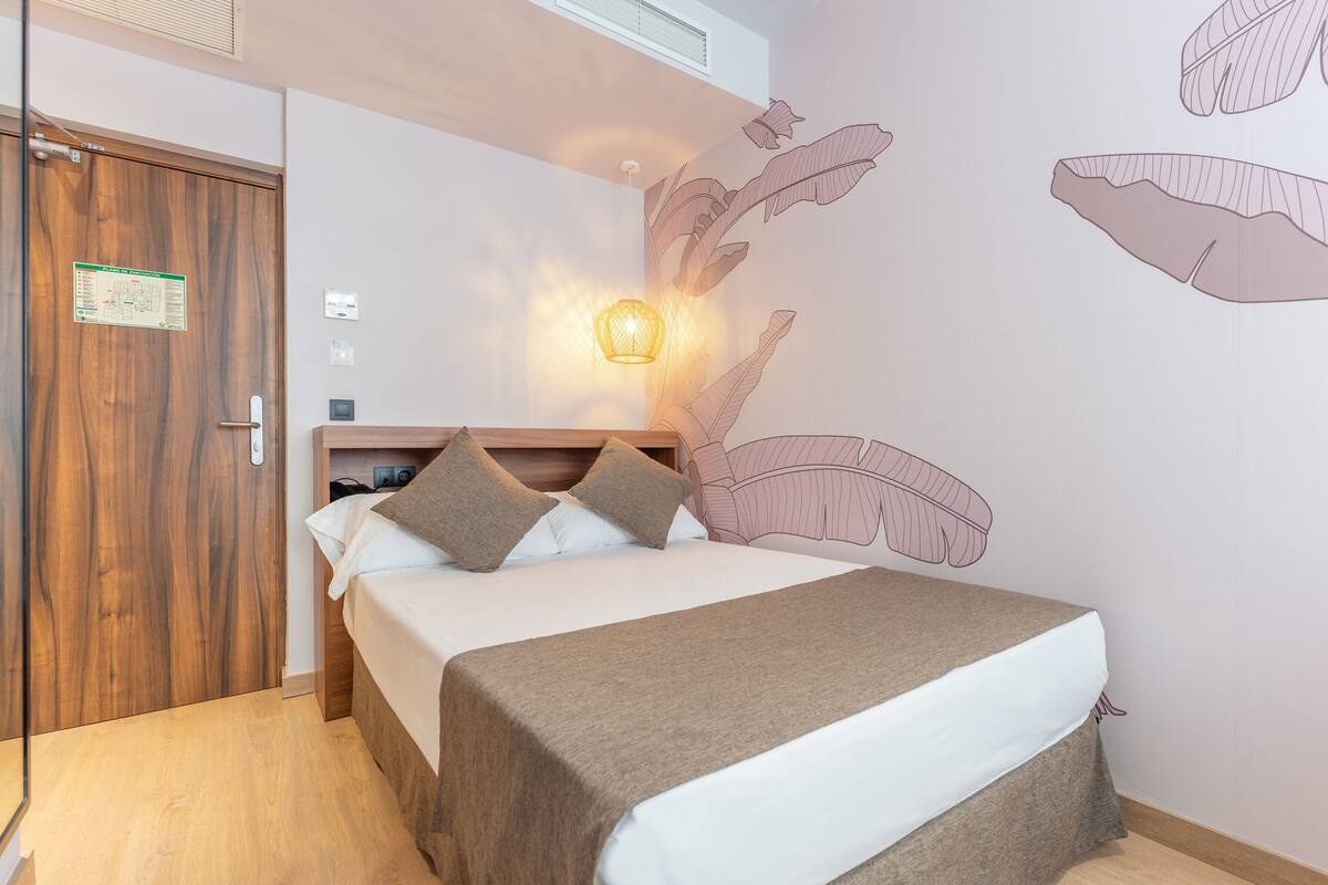 A bedroom with a wooden door, a bed with a white and brown bedspread, and a wall with leaf patterns.
