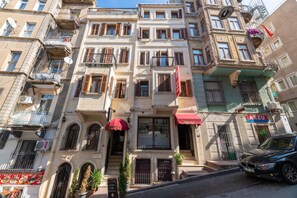 Un edificio a più piani con un hotel al piano terra, con balconi, un tendone rosso e un’auto parcheggiata.