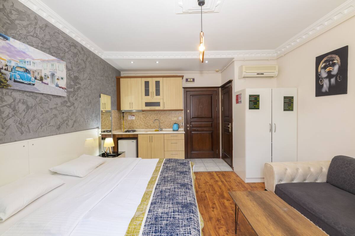 A hotel room with a bed, a sofa, a wooden bench, a kitchen area, and a bathroom.