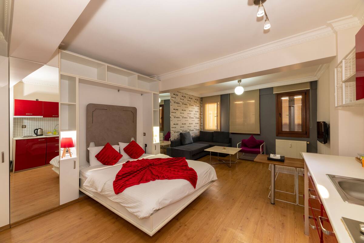 A modern hotel room with a bed, a sofa, a small dining table, and a kitchenette.