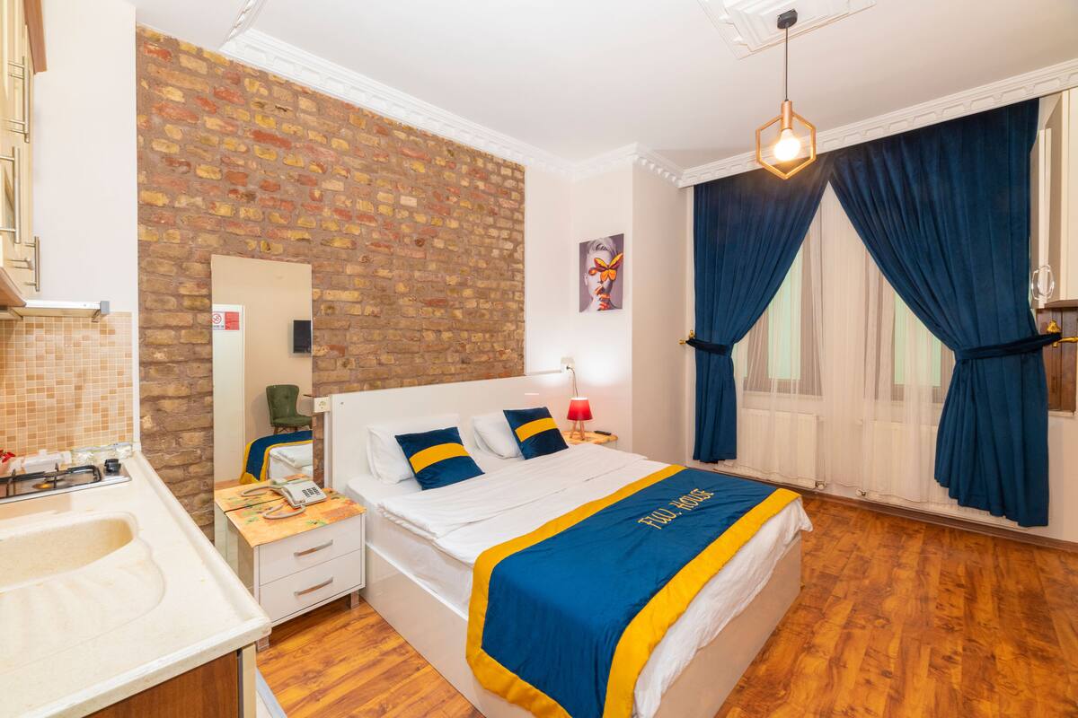 A hotel room with a brick wall, a bed with a blue and yellow bedspread, a wooden floor, a white bedside table, a red lamp, and a window with blue curtains.