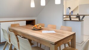 Et spiseområde med et træbord, seks stole og en skål med frugt. Tre hængende lamper er placeret over bordet.